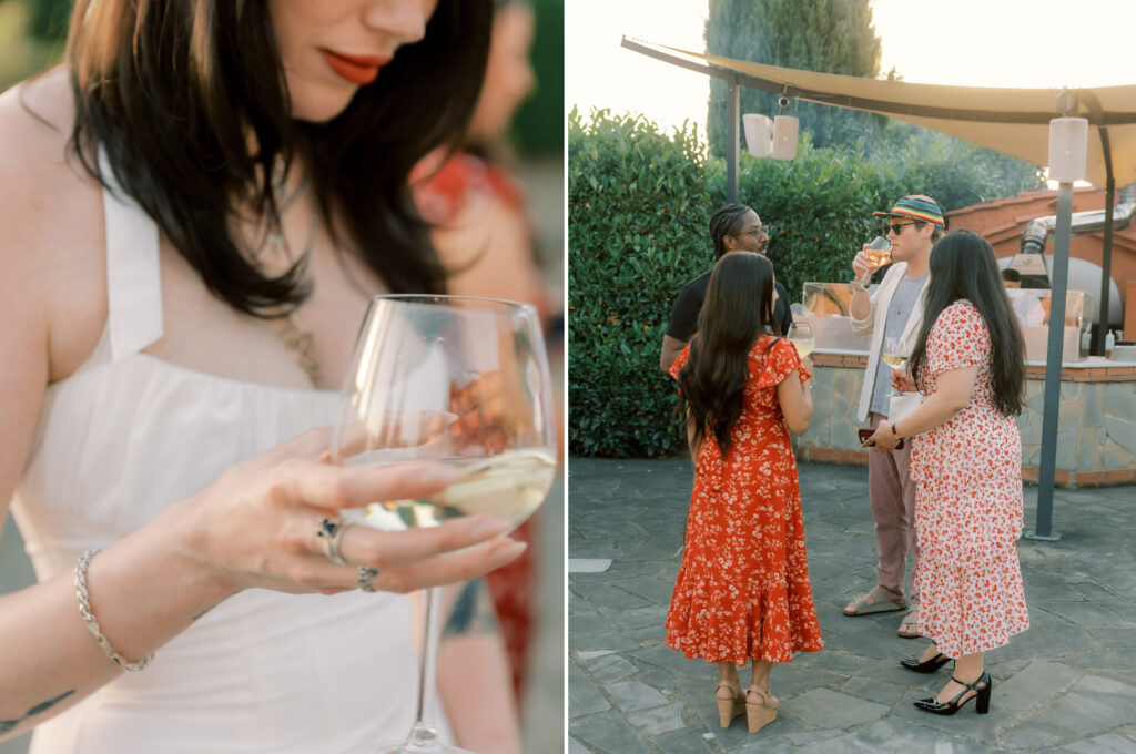 A side-by-side diptych of the bride and wedding guests at an outdoor Tuscan reception at golden hour. On the left, a close-up of the bride — a dark-haired woman in a white dress — holding a large glass of white wine, her silver bracelet and rings visible, with a soft smile and red lips. On the right, a wider shot on a stone terrace shows four guests chatting — two women in red floral midi dresses and a man in a colorful striped hat sipping wine — with a lush green hedge, cypress trees, and a canopied outdoor bar structure in the warm evening light.