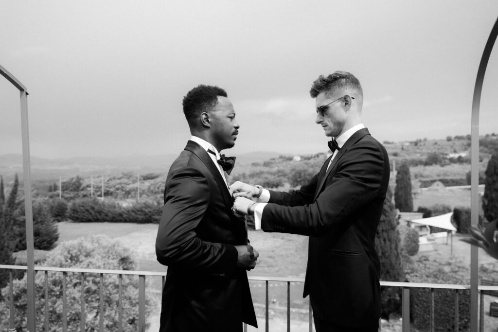 A black-and-white photograph of the groom standing on the villa terrace balcony while a groomsman in a matching black tuxedo carefully pins the boutonniere onto his lapel, both looking down with focus and quiet camaraderie. Rolling Tuscan hills, cypress trees, and the estate grounds stretch across the horizon behind them.