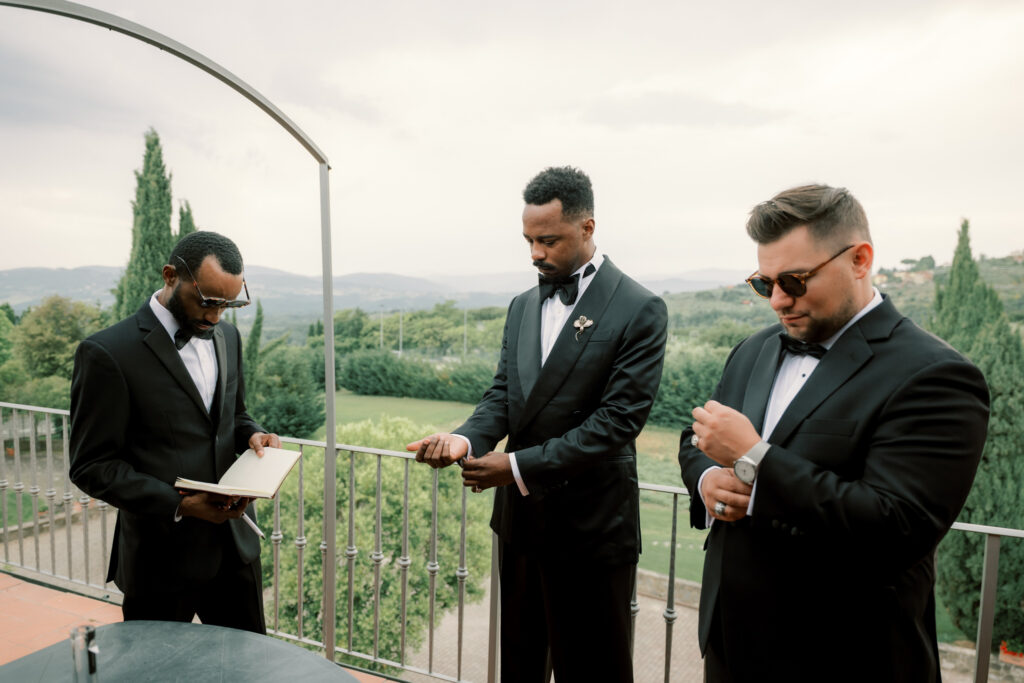 The groom and two groomsmen in black tuxedos and bow ties prepare on a terrace balcony at the Tuscan estate before the ceremony. One groomsman reads from a small book while the groom looks on, and another adjusts his cufflinks, with rolling Tuscan hills, cypress trees, and an overcast sky stretching behind them.