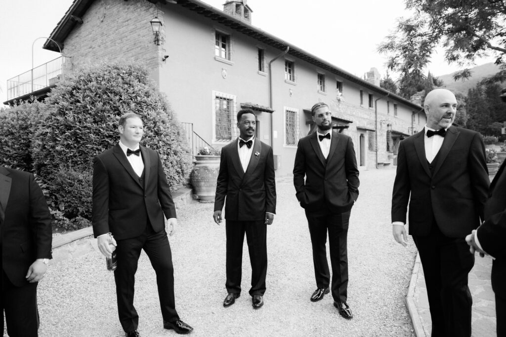 A black-and-white photograph of the groom and three groomsmen in matching black tuxedos and bow ties walking confidently along a gravel path in front of the rustic stone Tuscan villa facade, with terracotta pots and a stone staircase visible behind them.