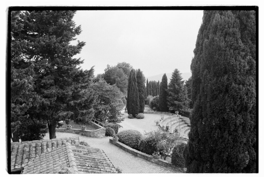 A black-and-white elevated view of a formal Italian garden, featuring a row of tall slender cypress trees leading toward distant hills, rounded topiary shrubs, stone retaining walls with potted plants, a curved stone staircase, and terracotta roof tiles in the foreground.