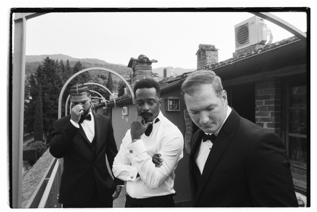 A candid black-and-white film photograph of three groomsmen in black tuxedos and bow ties gathered closely together on the villa terrace, in a relaxed and playful pre-ceremony moment with the Tuscan hills and rooftops visible behind them.