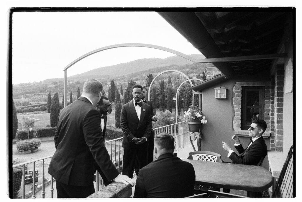 A wide black-and-white film photograph of the groom and groomsmen in black tuxedos relaxing on a stone terrace balcony before the ceremony, with some standing and conversing while one sits at an outdoor table smoking. A sweeping view of cypress trees, manicured gardens, rolling Tuscan hills, and a metal arch structure fills the dramatic background below.