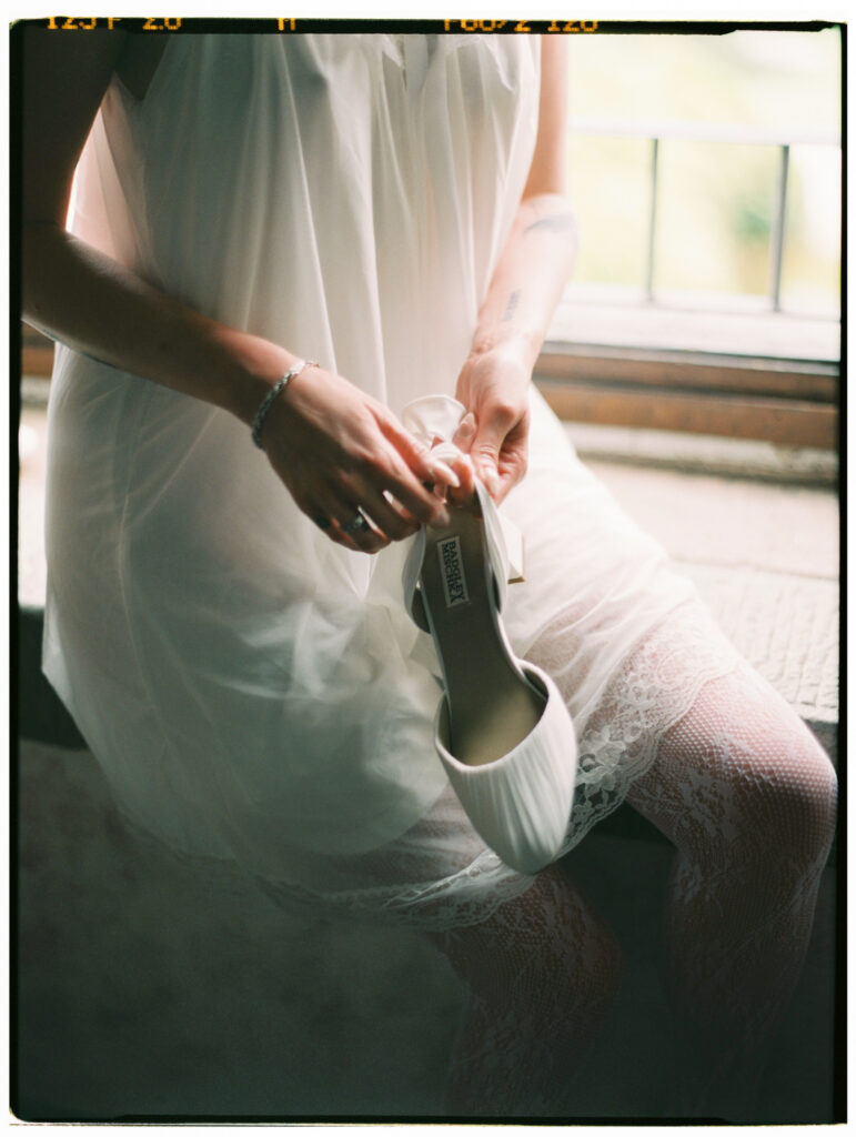 The bride sits perched on a wide stone windowsill in a white slip dress and lace stockings, fastening her wedding shoes, framed by tall arched French doors thrown open to a lush green Tuscan garden. Shot on film with a warm, dreamy quality.