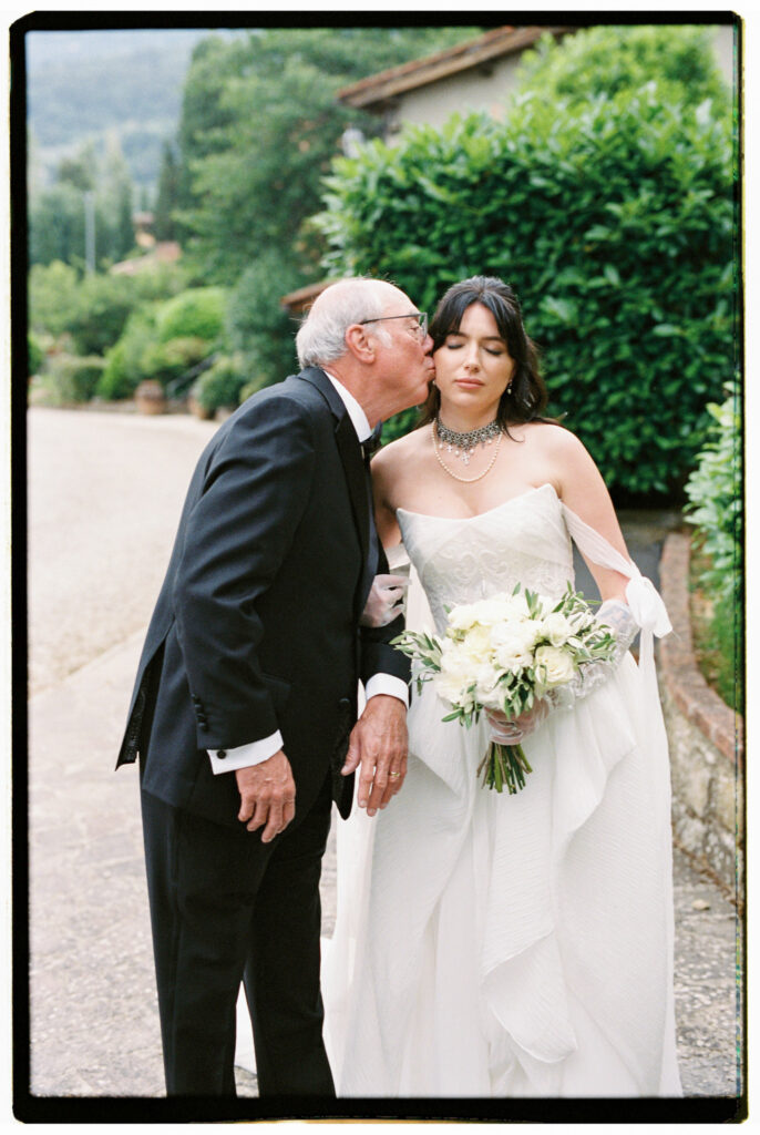 A tender film photograph of the bride in her off-the-shoulder white gown, crystal choker necklace, and long white gloves holding her white peony bouquet, receiving a kiss on the cheek from her father — in a black suit, outside the Tuscan estate amid lush green hedges.
