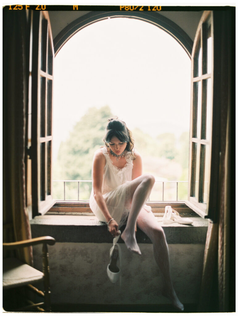 A film photograph of the bride sitting on a wide stone windowsill inside the Tuscan villa, wearing a delicate white slip dress, lace stockings, and a statement necklace, as she fastens her wedding shoes. She is framed by a tall arched window thrown open to a lush green garden, with warm, hazy light pouring in behind her and a vintage wooden chair visible to her left.