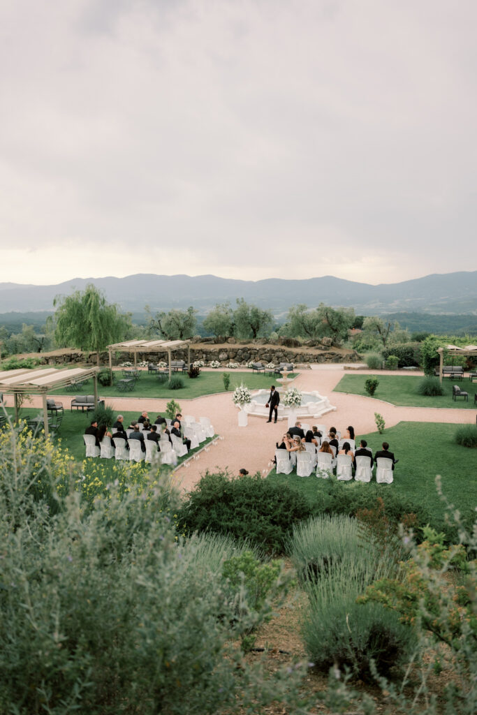 An elevated wide shot of an intimate outdoor wedding ceremony at a Tuscan estate, with guests seated in two small sections of white chair-covered seats on either side of a gravel path leading to a stone fountain altar adorned with tall white floral arrangements. A solitary figure stands at the altar awaiting the bride, surrounded by sweeping manicured lawns, weeping willow trees, lavender bushes, pergolas, and a dramatic backdrop of rolling mountain hills under a moody overcast sky.