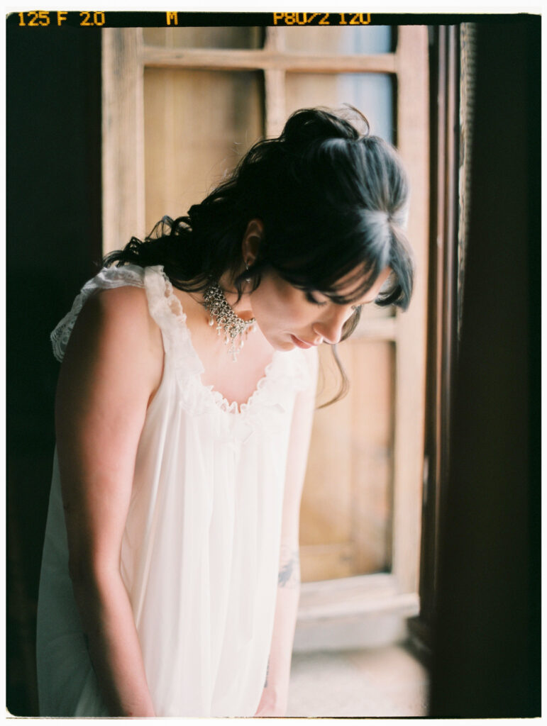 A intimate film photograph of the bride in a soft white lace-trimmed slip dress, her dark hair swept up in a loose updo with face-framing tendrils, gazing downward in a quiet, reflective moment beside a weathered wooden-framed window inside the Tuscan villa. A sparkling crystal chandelier earring and delicate lace shoulder straps are visible in the warm, moody light.