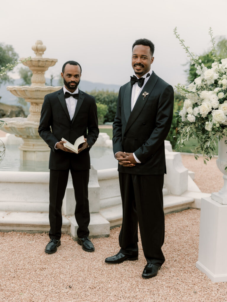 The groom stands at the outdoor altar beside the stone fountain, smiling warmly alongside the officiant who holds an open book, both in black tuxedos with the tall white floral arrangements and gravel ceremony grounds of the Tuscan estate behind them.