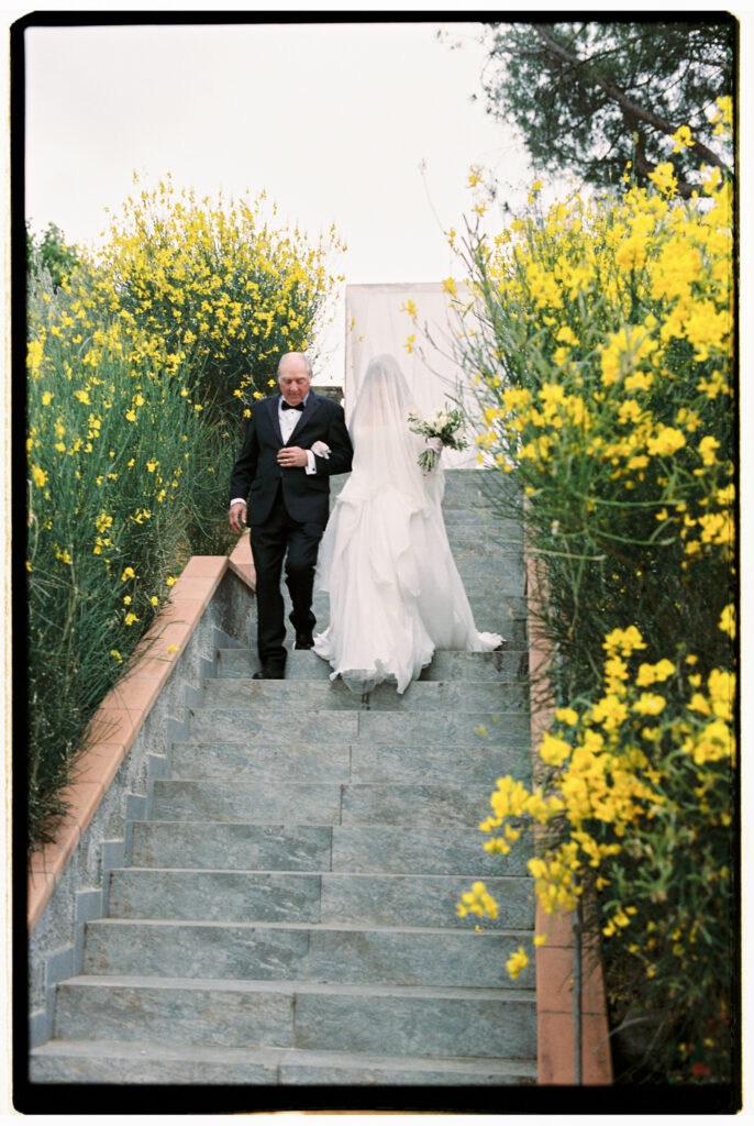 A film photograph of the bride descending wide stone steps on the arm of an older gentleman in a black tuxedo as she makes her processional entrance, her long cathedral veil trailing behind her, flanked by vibrant yellow wildflowers blooming on either side of the staircase.