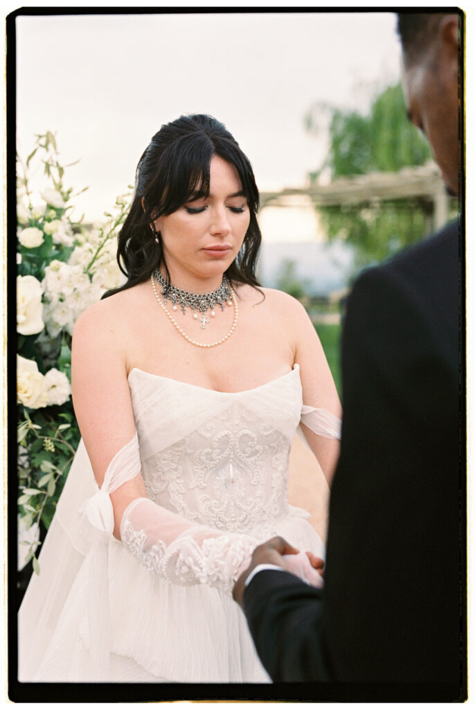 A close-up film portrait of the bride looking downward with a serene expression during the ceremony, wearing her off-the-shoulder lace corset gown, crystal and pearl choker necklace, and sheer white gloves, with white floral arrangements and a weeping willow softly blurred in the background.