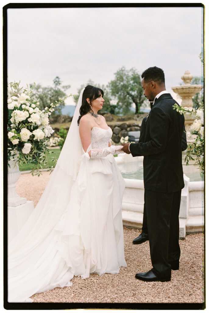 The bride and groom stand facing each other at the outdoor altar beside the stone fountain, exchanging vows during their intimate Tuscan wedding ceremony, surrounded by lush white floral arrangements, with the estate garden and olive trees softly visible behind them.
