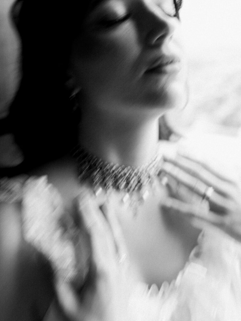 A dreamy, intentionally blurred black-and-white close-up of the bride fastening a multi-strand crystal or pearl choker necklace at her throat, her chin tilted upward and eyes closed. The motion blur and high contrast create an ethereal, editorial mood against a bright, overexposed background.