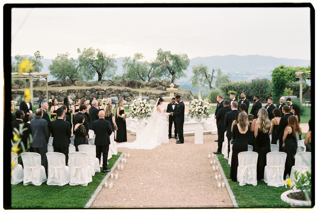 A sweeping film photograph of the full outdoor wedding ceremony at the Tuscan estate, with the bride in her white gown and cathedral veil and the groom in his black tuxedo exchanging vows at the stone fountain altar flanked by towering white floral arrangements. Guests dressed entirely in black formal attire stand on either side of a candle-lined gravel aisle with white slipcovered chairs, while the wedding party looks on. Ancient olive trees, a rugged stone wall, rolling mountain hills, and an overcast sky form a dramatic natural backdrop behind the couple.