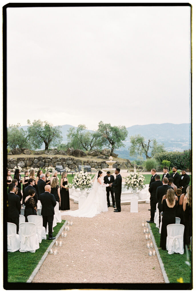 A wide ceremony shot looking down the aisle toward the altar, with candles lining the white runner, guests seated on white chair-covered seats on either side, the wedding party standing at the fountain altar adorned with white florals, and the sweeping Tuscan hillside and olive tree grove stretching into the distance under an overcast sky.