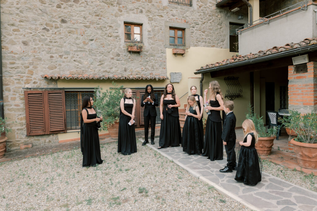 The wedding party gathers in the rustic cobblestone courtyard of the Tuscan stone villa before the ceremony. Six bridesmaids in elegant floor-length black gowns stand in a loose group receiving instructions, joined by a flower girl and a young page boy also dressed in black, with terracotta pots, wooden shuttered windows, and the textured stone facade of the villa behind them.