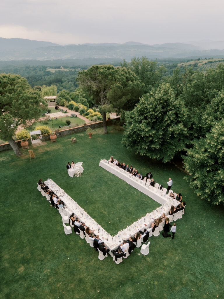 A dramatic aerial drone shot of the outdoor wedding reception on the Tuscan estate lawn, showing the U-shaped arrangement of long white linen banquet tables filled with guests dressed in black formal attire, surrounded by lush green trees, yellow wildflowers, and sweeping mountain views beyond the stone perimeter wall.