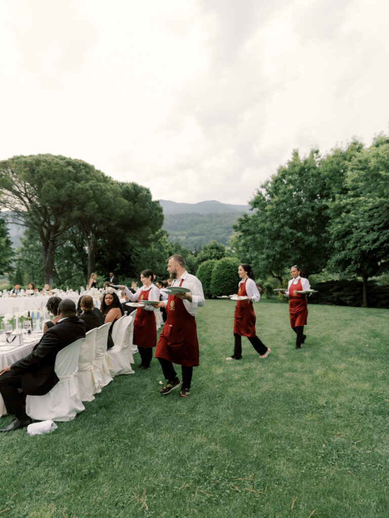 Three servers in burgundy aprons carry large silver platters across the green lawn to waiting guests at the outdoor banquet tables, with the Tuscan hillside, stone pine trees, and an overcast sky visible behind them.