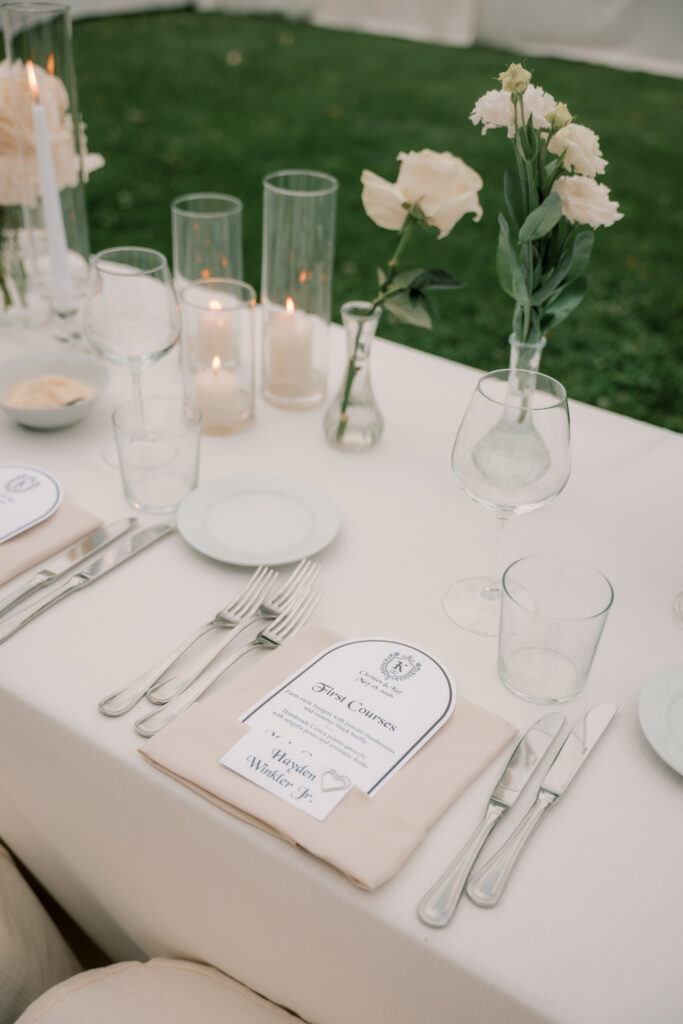 A close-up detail shot of an elegantly styled place setting at the outdoor reception, featuring silver cutlery, a folded linen napkin, a printed menu card, tall glass hurricane candle holders, and a single white flower in a glass bud vase on a white linen tablecloth.