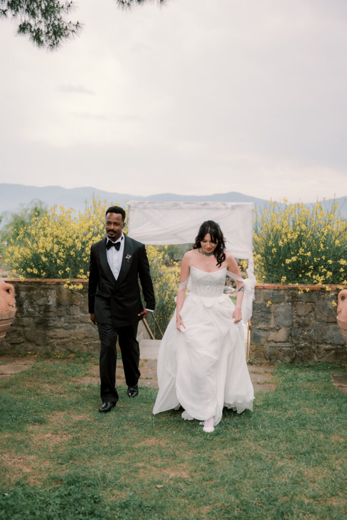 The bride and groom make their grand entrance into the outdoor reception on the Tuscan estate lawn, walking hand in hand and smiling, with yellow wildflowers, a stone terrace wall, and the rolling Tuscan mountain panorama glowing behind them under a soft overcast sky.