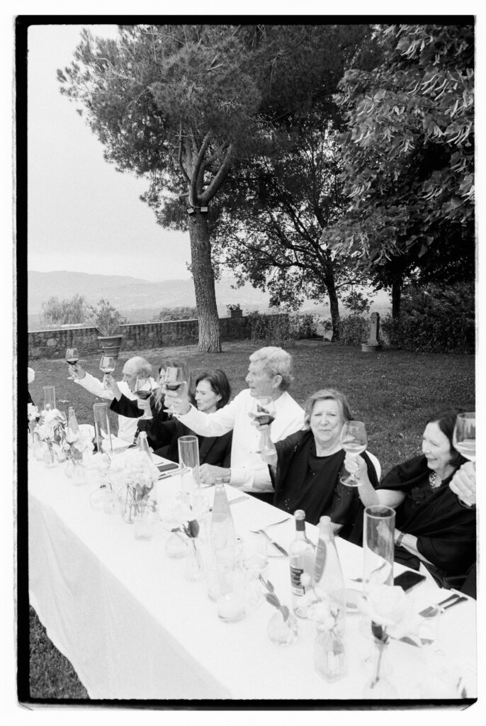A candid black-and-white film photograph of female guests at the banquet table raising their wine glasses in a joyful toast, laughing together beneath a large stone pine tree with the Tuscan countryside stretching into the background.