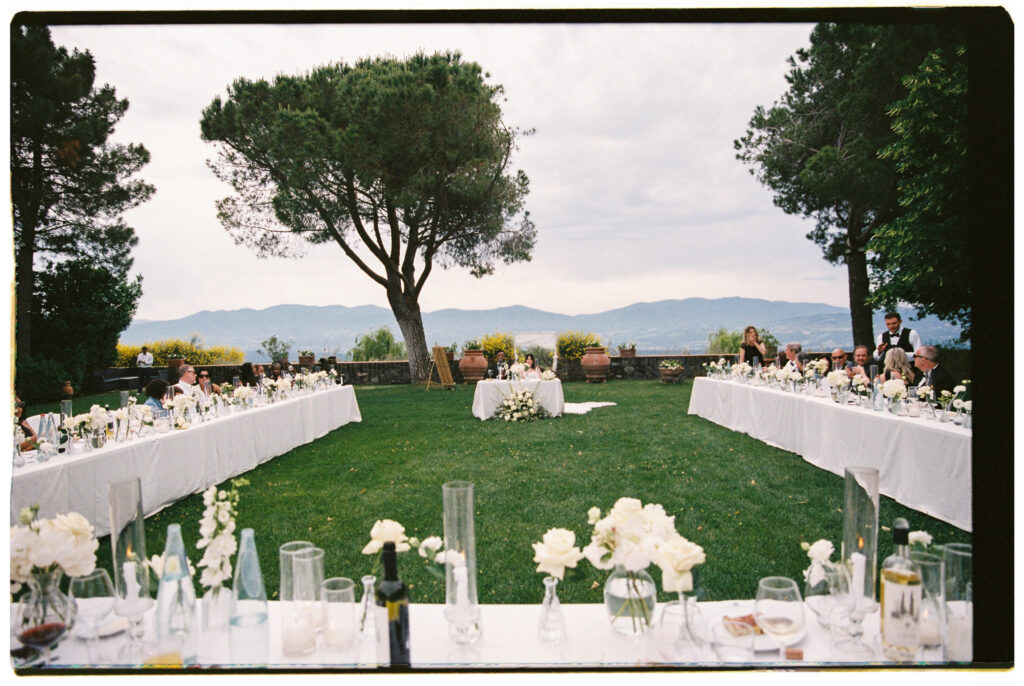 A film photograph looking down the center of an elegant outdoor wedding reception on a lush green lawn at the Tuscan estate, with two long white linen-draped banquet tables lined with guests in formal black attire on either side. The tables are styled with clusters of white roses, tall glass bud vases, wine bottles, and glassware. The bride and groom sit at a small sweetheart table at the far end, framed by a large Italian stone pine tree, terracotta urns, yellow flowering shrubs, and a breathtaking panoramic view of the rolling Tuscan mountain range under a dramatic cloudy sky.