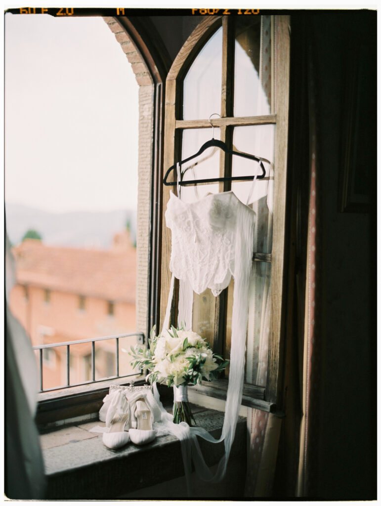 A film photograph of the bride's white lace wedding gown hanging from a hanger on an arched gothic-style window inside the Tuscan villa, with her white bridal heels, a bouquet of white flowers, and a flowing veil arranged on the stone windowsill, the Tuscan rooftops and countryside visible through the glass behind.
