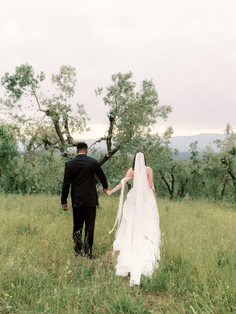 A bride and groom seen from behind, walking hand in hand through a wild green meadow toward olive trees and rolling Tuscan hills under a soft, overcast sky. The bride's long white veil and gown trail behind her as the groom, dressed in a black suit, leads her forward.
