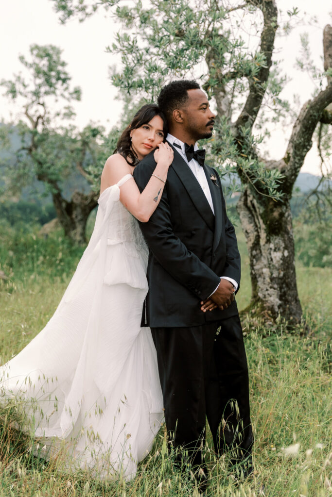 A bride in an off-the-shoulder white tiered wedding gown rests her cheek against the shoulder of a groom dressed in a black tuxedo with a bow tie, as they stand among olive trees and tall grass in the Tuscan countryside.