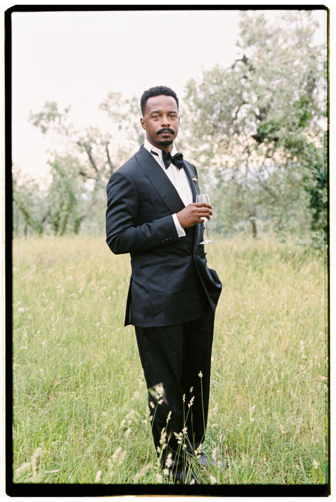 The groom stands confidently in a wild green Tuscan meadow, holding a champagne flute and gazing directly at the camera, dressed in a black tuxedo with a bow tie and white dress shirt, with flowering trees in the soft background.
