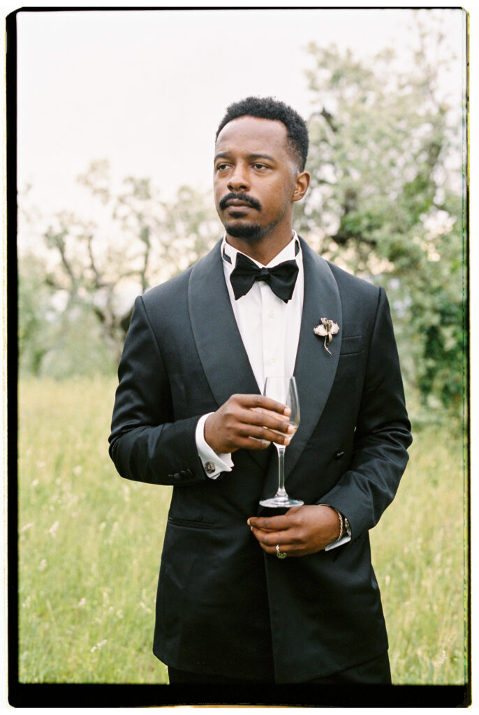 A film portrait of the groom in his black tuxedo and bow tie holding a champagne flute, gazing pensively into the distance against a lush green Tuscan meadow with flowering olive trees behind him.