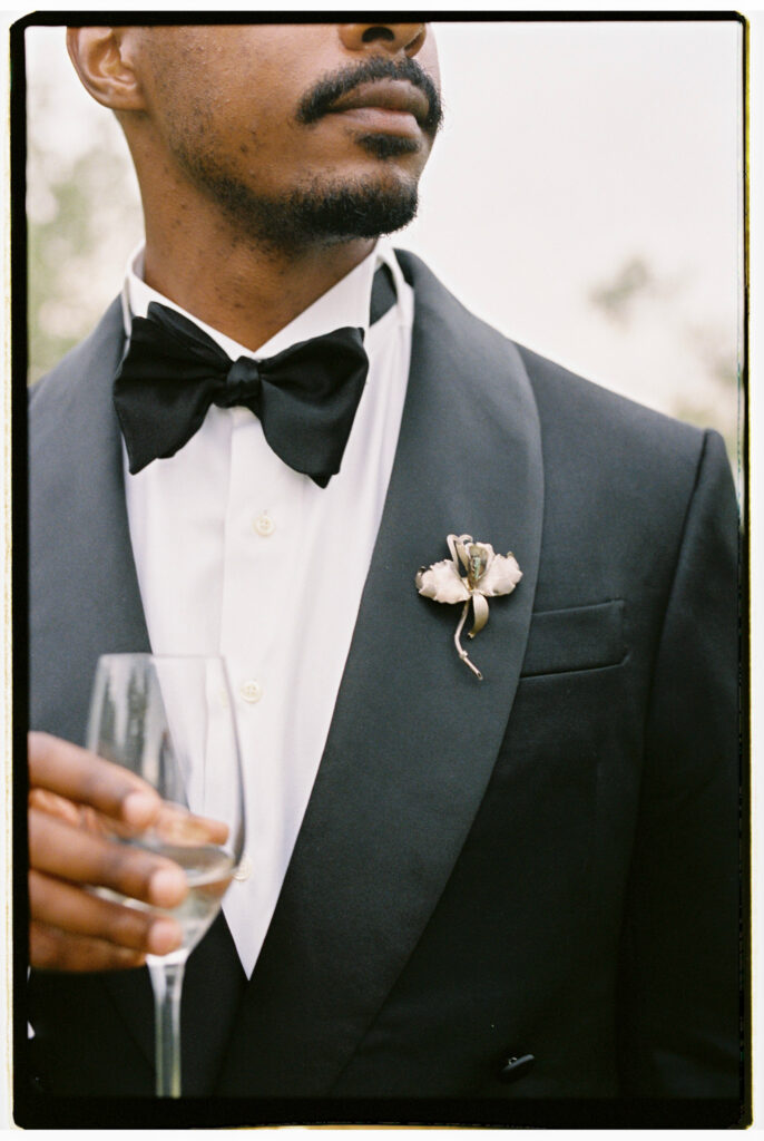 A close-up film detail shot of the groom's black tuxedo lapel, featuring a delicate floral boutonniere, a crisp white dress shirt, black bow tie, and a champagne flute held at his side.