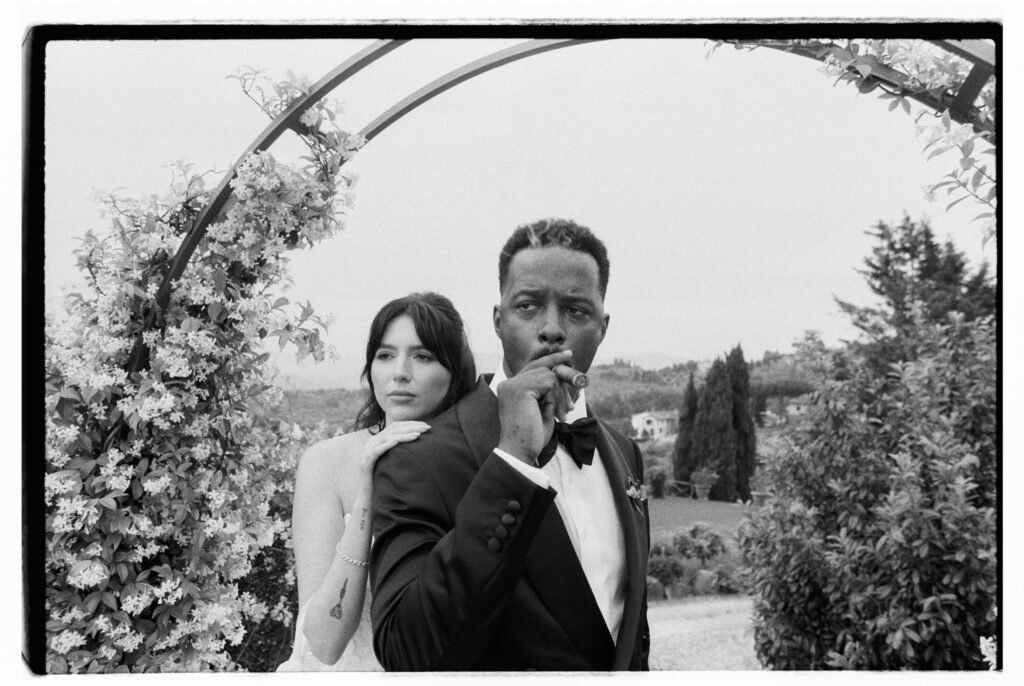 A striking black-and-white film photograph of the bride and groom standing beneath a floral arch draped in blooming flowers at the Tuscan estate. The bride leans against the groom's back, her hand resting on his shoulder and forearm tattoo visible, gazing coolly at the camera, while the groom smokes a cigar and stares directly into the lens with quiet confidence. Rolling Tuscan hills, cypress trees, and manicured hedges stretch behind them.