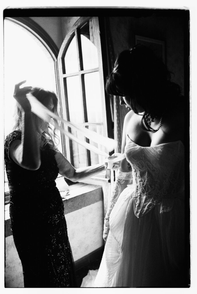 A moody black-and-white film photograph of the bride being helped into her wedding dress by an attendant near a tall arched window inside the Tuscan villa, dramatic light casting deep shadows across the intimate getting-ready moment.