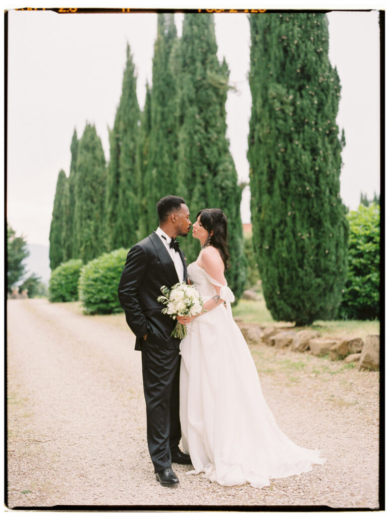 The bride and groom share a kiss on a tree-lined gravel driveway at the Tuscan estate, flanked by tall cypress trees and manicured hedges. The bride holds her white peony and olive branch bouquet while her flowing gown and train spread across the path.