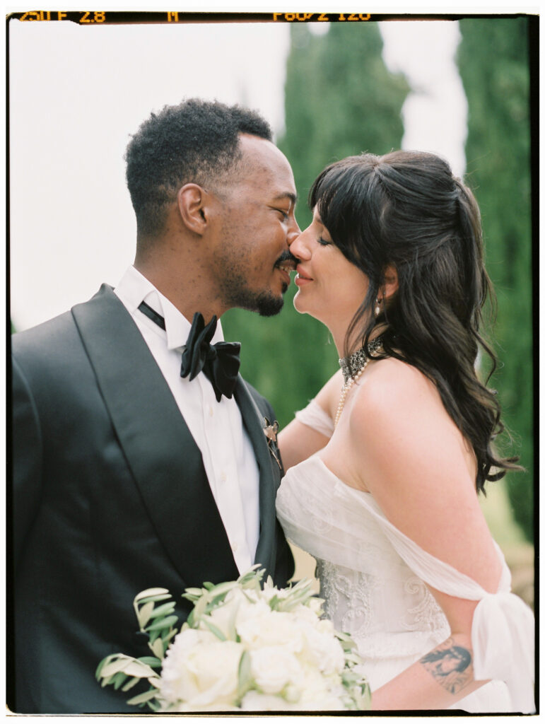A tender close-up film photograph of the bride and groom leaning in for a kiss, foreheads nearly touching, with the bride in her off-the-shoulder lace gown and crystal choker necklace and the groom in his black tuxedo and bow tie.
