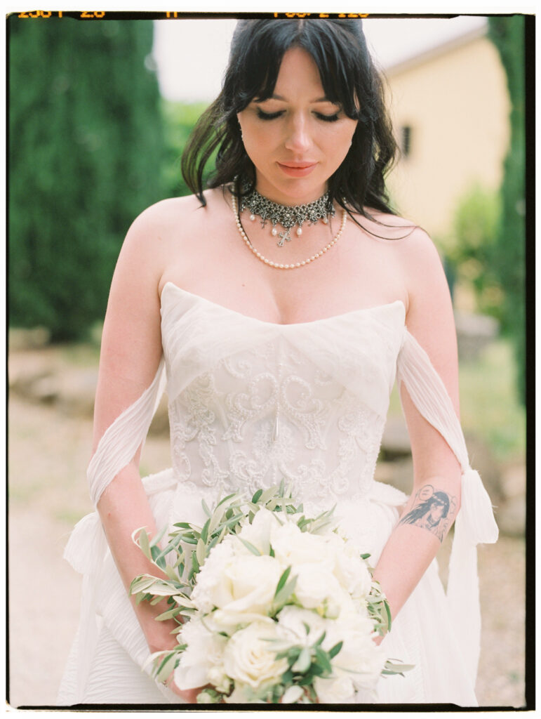 A close-up film portrait of the bride looking downward with eyes closed, wearing an off-the-shoulder white lace corset wedding gown and layered bridal jewelry — an ornate crystal and pearl choker with a cross pendant over a delicate pearl strand necklace. She holds a loose bouquet of white peonies and olive branches, her forearm tattoo visible, with cypress trees and the soft-focus Tuscan estate in the background.
