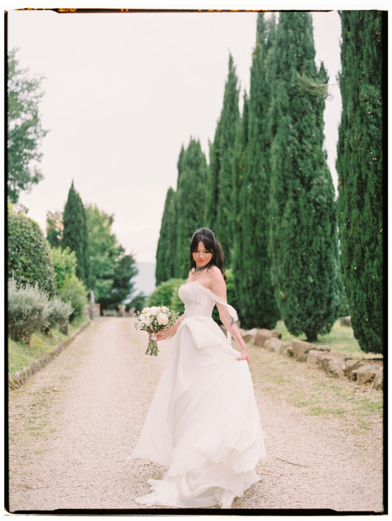 The bride stands alone on a grand cypress-lined gravel driveway at the Tuscan estate, her off-the-shoulder white gown and veil billowing softly, holding her bouquet as she looks back toward the camera with the row of towering cypress trees receding dramatically behind her.