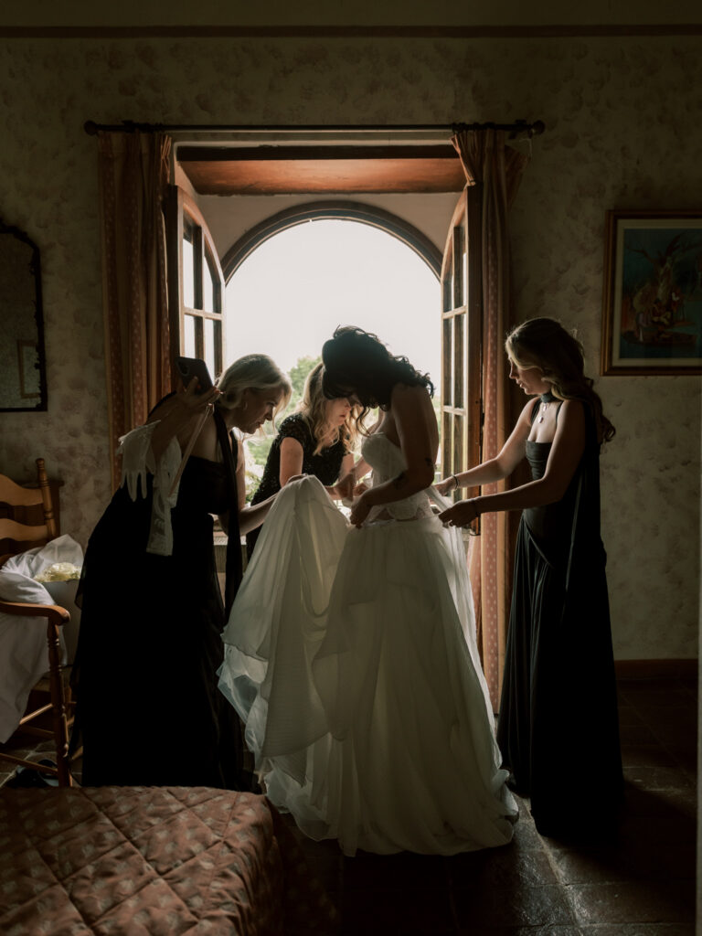 A beautifully backlit shot of the bride in her full white wedding gown being attended to by two helpers in a warmly lit villa room, framed by a grand arched doorway that floods the scene with soft natural light from the Tuscan garden beyond.