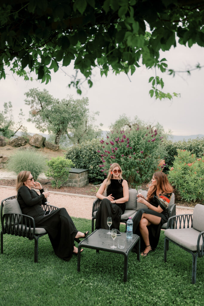 Three female wedding guests in elegant black dresses relax on outdoor lounge chairs beneath a shady tree in the Tuscan estate gardens, chatting and enjoying drinks on a glass side table, with rolling hills and flowering plants visible in the soft background.