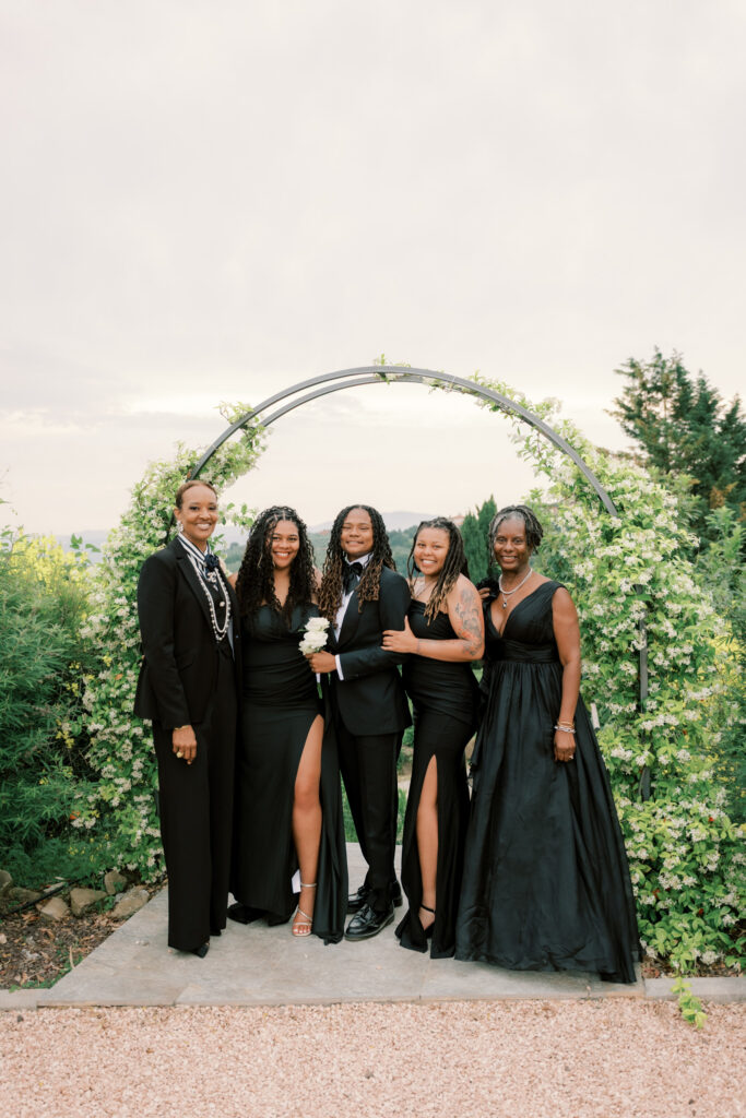 Four guests — two men in black tuxedos and two women in elegant black gowns — pose together smiling beneath the floral arch at the Tuscan estate, with the overcast sky and lush greenery of the garden behind them.