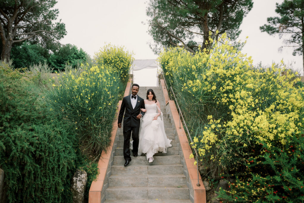 The bride and groom walk arm in arm down stone garden steps at the Tuscan estate, both laughing joyfully, surrounded by wild yellow flowering plants. The bride gathers her flowing white gown as the groom in his black tuxedo guides her down the path.