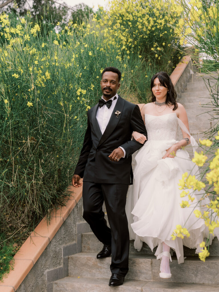 The bride and groom walk arm in arm down stone garden steps at the Tuscan estate, both laughing joyfully, surrounded by wild yellow flowering plants. The bride gathers her flowing white gown as the groom in his black tuxedo guides her down the path.