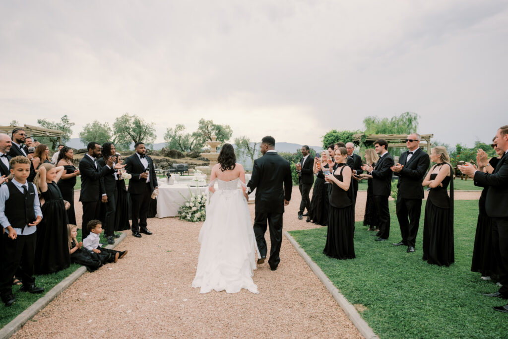 A wide shot of the bride and groom seen from behind, walking hand in hand down a gravel garden path as their wedding guests — all dressed in black formal attire — line both sides, clapping and cheering in celebration. The Tuscan countryside, weeping willow trees, and an overcast sky stretch across the horizon in the background.