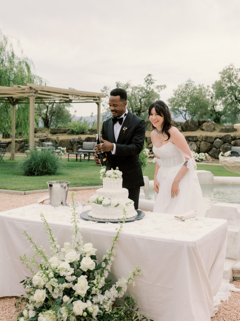 The bride and groom stand together at an elegantly dressed outdoor cake table, the groom popping a bottle of champagne beside a tall white tiered wedding cake adorned with white floral arrangements and greenery, set against the lush Tuscan garden and pergola in the background.