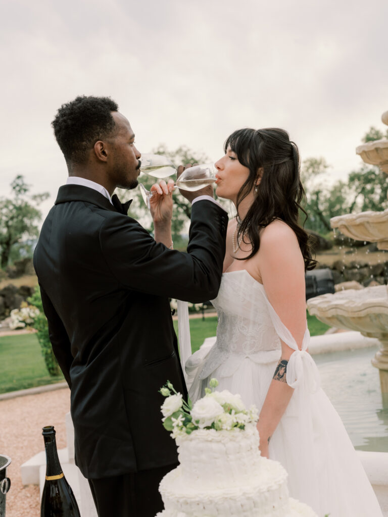 A candid close-up of the groom feeding the bride a sip of champagne directly from a glass at the cake table, both sharing a playful, intimate moment beside the white tiered wedding cake, with the Tuscan estate grounds visible behind them.