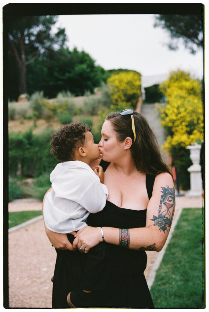 A candid film photograph of a female wedding guest in a black sleeveless dress, her arms tattooed, kissing a young toddler boy dressed in a white shirt on the cheek in the lush green gardens of the Tuscan estate.