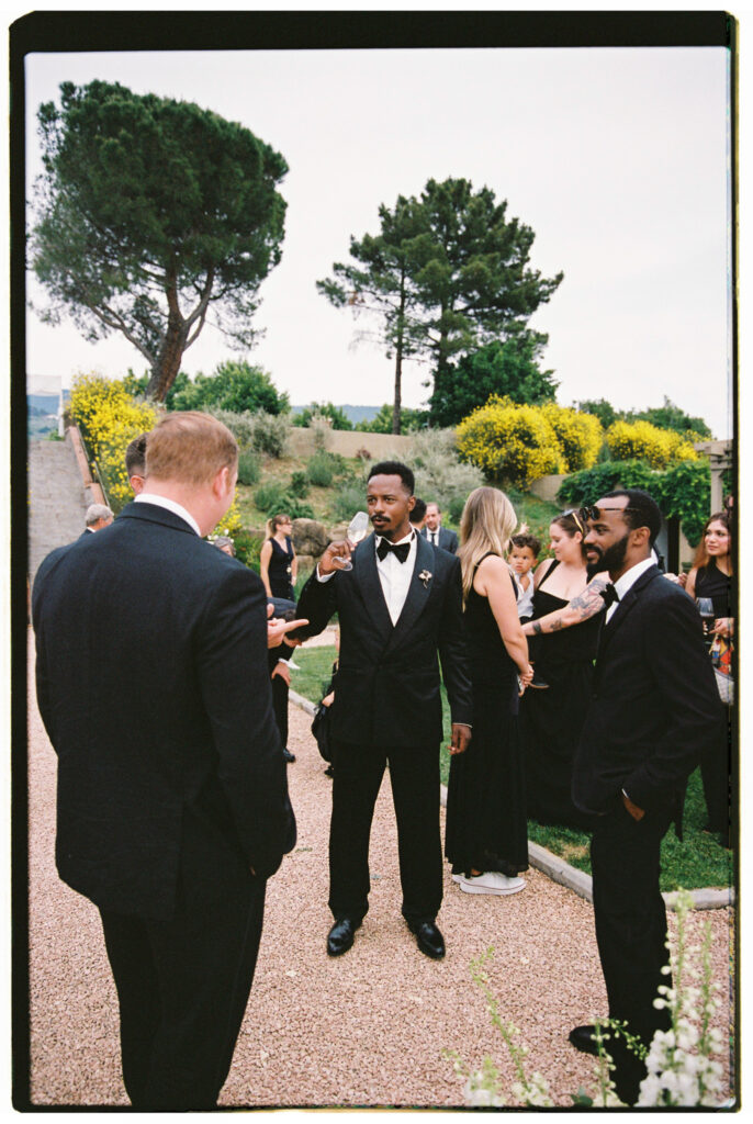 A film photograph of the groom in his black tuxedo and bow tie greeting guests on a gravel garden path at the Tuscan estate, smiling warmly as he shakes hands, with other wedding guests in formal black attire mingling in the background among flowering yellow shrubs and tall pine trees.
