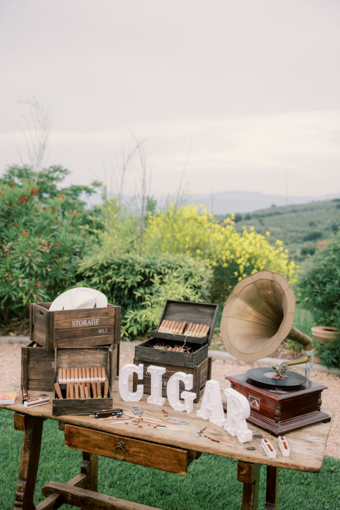 A styled cigar station set up on a rustic wooden table at the outdoor wedding, featuring an open wooden cigar box, individual cigars laid out, a vintage brass gramophone, and a marquee letter sign reading "CIGAR," set against a backdrop of yellow wildflowers and the Tuscan countryside.