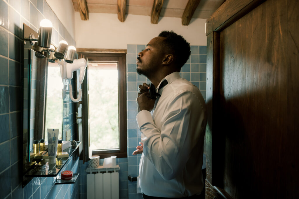The groom stands in profile in a rustic Tuscan villa bathroom with blue tiled walls and exposed wooden ceiling beams, fastening his black bow tie over a crisp white dress shirt as he gets ready on his wedding day. Warm light streams through a wooden-framed window, illuminating grooming products on the vanity shelf beside a lit mirror.